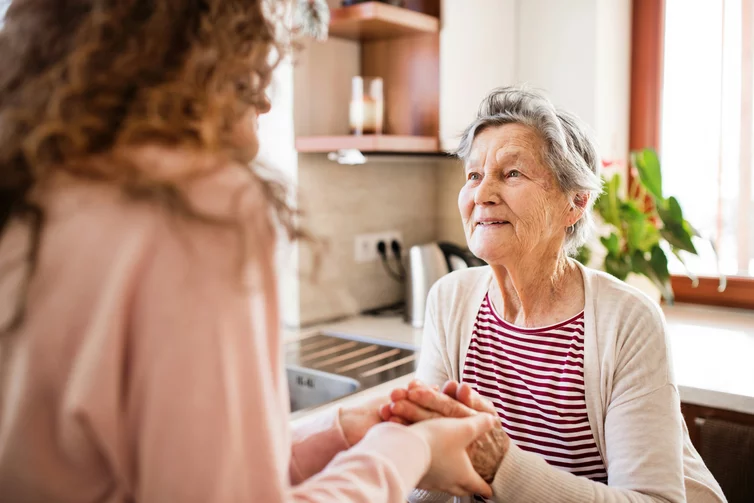 Eine junge Frau hält die gefalteten Hände einer sie anschauenden Seniorin in den eigenen Händen.
