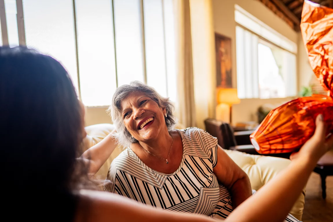Eine junge Frau und eine Seniorin sitzen gemeinsam auf einem Sofa. Die junge Frau schaut die lächelnde Seniorin an. iStock.com/FG Trade