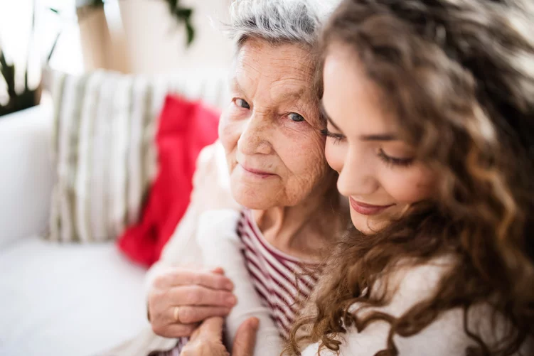 Eine junge Frau umarmt innig mit geschlossenen Augen von hinten eine Seniorin. Die Seniorin schaut friedlich in die Kamera. iStock.com/Halfpoint