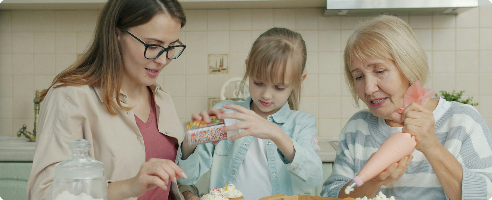 Drei Generationen von Frauen: eine Seniorin, eine Erwachsene und ein Mädchen backen gemeinsam Cup-Cakes in einer Küche.