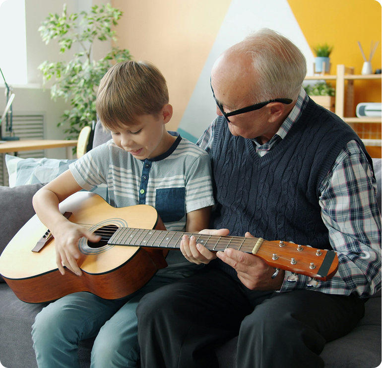 Ein senioriger Mann und ein Junge spielen Gemeinsam auf einer Gitarre. Der Junge hat die Gitarre dabei auf dem Schoß, während der ältere Mann sanft den Hals der Gitarre hält.