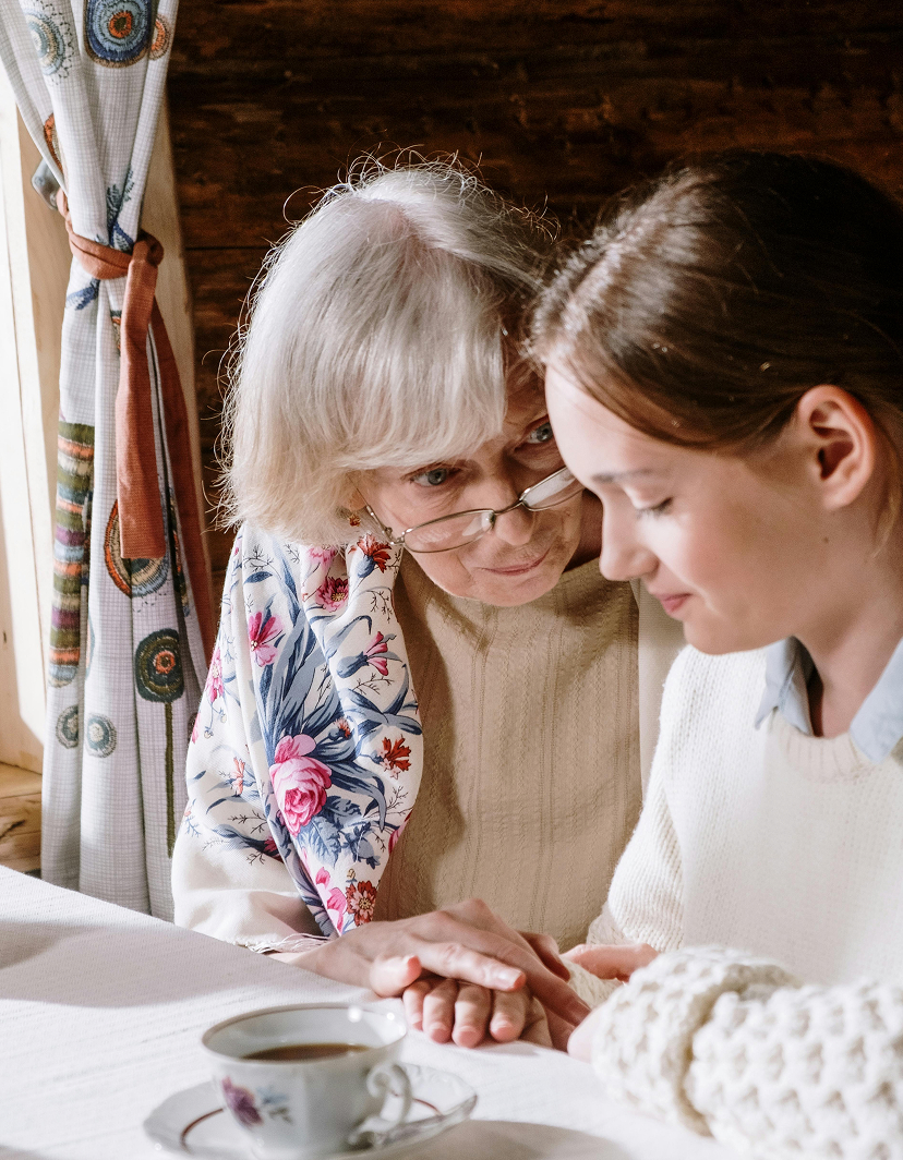 Eine Seniorin sitzt gemeinsam mit einem Mädchen am Tisch und hält die Hände des Mädchens behutsam in ihren eigenen. Die Seniorin schaut das Mädchen ihr zugeneigt über ihre Brille hinweg an während das Mädchen auf den Tisch schaut. 