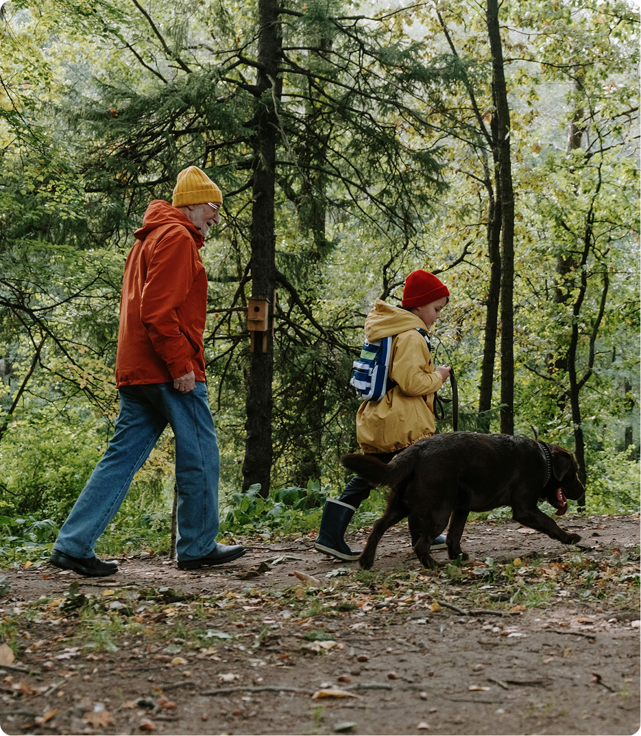 Ein Senioriger Mann mit Wanderstock und ein Junge spazieren gemeinsam mit einem Hund einen Waldweg entlang.
