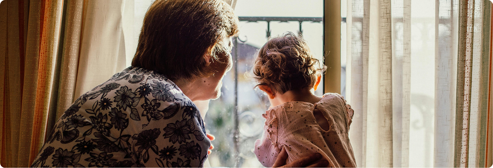 Eine Seniorin und ein Kleinkind schauen gemeinsam aus dem Fenster. Die Seniorin lächelt nach Draußen.